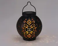 Rund sort lanterne med indviklede udskæringer, der lader varmt gult lys skinne igennem og skaber dekorative lysmønstre på omgivelserne. Lanternen har et håndtag øverst til nem transport eller ophængning samt en flad top, der muligvis rummer et solpanel til udendørs brug.