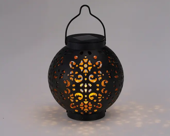Rund sort lanterne med indviklede udskæringer, der lader varmt gult lys skinne igennem og skaber dekorative lysmønstre på omgivelserne. Lanternen har et håndtag øverst til nem transport eller ophængning samt en flad top, der muligvis rummer et solpanel til udendørs brug.