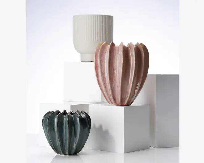 Tre dekorative beholdere vises, hver med et unikt design. Foran til venstre er en mørkegrøn beholder med blank overflade og bølget, græskar-lignende form. I midten står en højere, lyserød beholder med skinnende glasur og dybe lodrette riller, også med et stiliseret græskarudtryk. Bagest er en hvid, cylindrisk beholder med mat finish og diskrete lodrette riller. Alle beholdere er lavet af keramik og har et moderne, kunstnerisk udtryk.