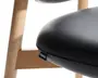 Nærbillede af et stole sæde med glat, sort kunstlæder polstring og en let tekstur. Sædet er tykt polstret for komfort og har en lille sort stofmærke fastgjort til sømmen. Stolens ramme er lavet af lyst træ med en naturlig finish, der viser rene linjer og solid konstruktion.