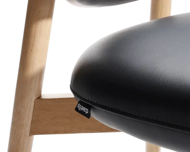 Nærbillede af et stole sæde med glat, sort kunstlæder polstring og en let tekstur. Sædet er tykt polstret for komfort og har en lille sort stofmærke fastgjort til sømmen. Stolens ramme er lavet af lyst træ med en naturlig finish, der viser rene linjer og solid konstruktion.