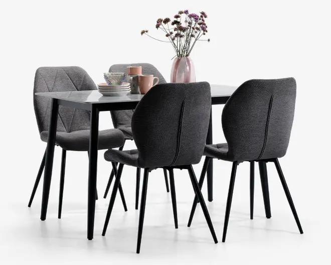 Et moderne spisesæt med et rektangulært bord, der har en glat, blank hvid bordplade og slanke sorte ben. Sættet inkluderer fire polstrede stole i mørkegråt stof, hver med buet ryglæn og lodrette syninger for ekstra komfort og stil. Stolene har slanke sorte metalben. Bordet er pyntet med en lyserød vase med lilla og hvide blomster samt et sæt stablede tallerkener, skåle og krus i afdæmpede pastelfarver.