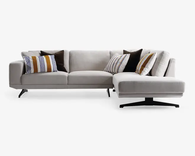 En moderne L-formet sofa med lyse beige betræk, rene linjer og lave metalben. Sofaen har flere puder: nogle er ensfarvede mørkebrune, mens andre har hvid baggrund med lodrette striber i sennepsgul, brun og sort. Sæde- og ryghynderne ser bløde og støttende ud, og sofaens højre side har en forlænget chaiselong.