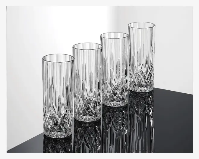 Sæt med fire høje, klare drikkeglas med facetslebet diamantmønster i bunden, der går over i lodrette riller langs siderne. Glassene har en cylindrisk form og en glat kant, og de reflekterer lyset, hvilket fremhæver det detaljerede design.