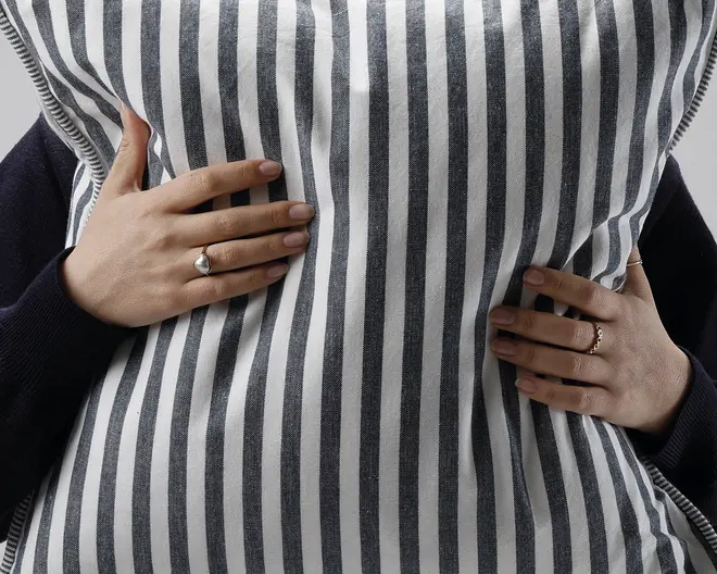 Nærbillede af en rektangulær pude med lodrette sorte og hvide striber. Stoffet ser blødt og let tekstureret ud. Puden bliver omfavnet af to hænder, der hver bærer en fin ring – den ene med en perlelignende sten og den anden med en tynd guldring. Pudens kanter har en diskret syet kant.