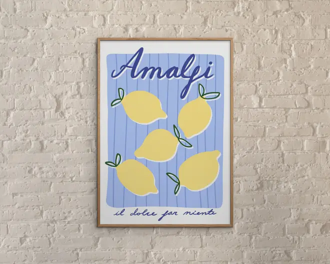 Indrammet kunsttryk med en legende illustration af fem gule citroner med grønne blade på en lyseblå baggrund med lodrette striber. Øverst står ordet 'Amalfi' med store, blå, kursiverede bogstaver, og nederst ses sætningen 'il dolce far niente' skrevet med håndskrift. Trykket har en moderne, minimalistisk stil og er vist mod en hvid murstensvæg med struktur.