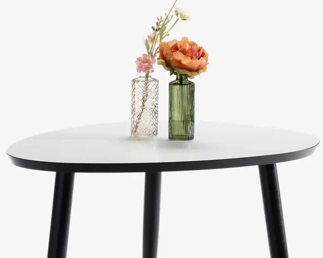 Ovalt bord med en glat, hvid bordplade og fire sorte, let skrå ben. Ovenpå står to dekorative vaser: den ene er klar med mønstret overflade og indeholder små lyserøde og hvide blomster, mens den anden er af grønt glas med lodrette riller og rummer en stor orange blomst samt nogle hvide blomster.