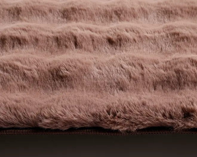 Nærbillede af et blødt, plysset stof med bølget, tekstureret overflade. Materialet virker tykt og fluffy i en lys brun eller taupe farve. De hævede riller giver et hyggeligt og luksuriøst udtryk, ideelt til varme og komfort.