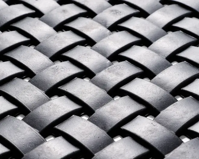 Nærbillede af et vævet mønster lavet af brede, mat sorte strimler. Strimlerne er flettet sammen i et over-under mønster, hvilket skaber en tekstureret, kurvelignende overflade. Materialet virker robust og let tekstureret med en diskret glans på overfladen.