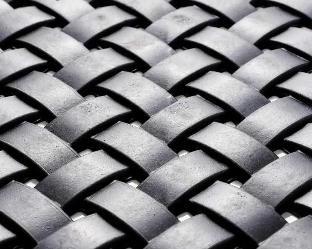 Nærbillede af et vævet mønster lavet af brede, mat sorte strimler. Strimlerne er flettet sammen i et over-under mønster, hvilket skaber en tekstureret, kurvelignende overflade. Materialet virker robust og let tekstureret med en diskret glans på overfladen.