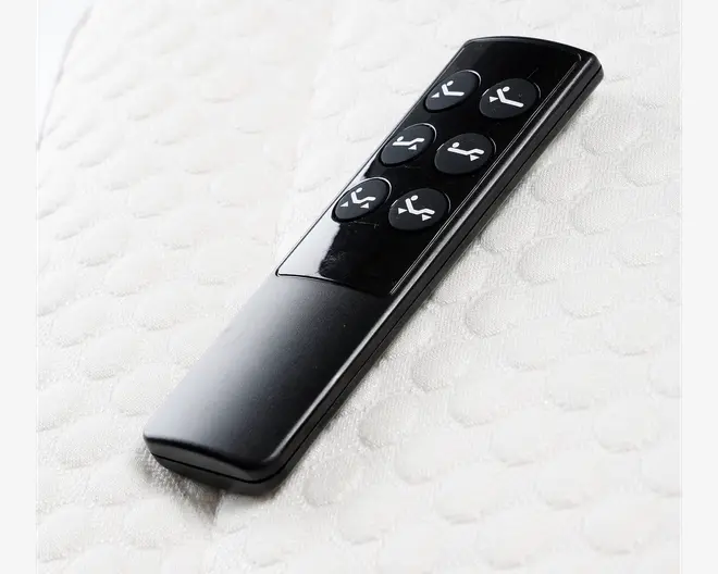 Elegant sort fjernbetjening med seks runde knapper, hver med hvide ikoner, der viser forskellige justerbare positioner til en seng. Knapperne er tydeligt markeret med enkle illustrationer for at hæve eller sænke hoved- og fodende samt en flad position. Fjernbetjeningen har en glat, blank overflade og en ergonomisk form, der ligger godt i hånden.