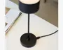 Nærbillede af en moderne, minimalistisk sort lampe med cylindrisk fod og slank, lodret stang. Lampen får strøm via et USB-kabel, der er tilsluttet basen. Overfladen er mat sort, og designet er stilrent og enkelt, velegnet til moderne indretning.