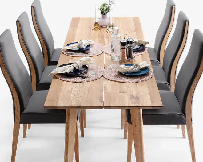 Rektangulært spisebord i træ med naturfinish, omgivet af seks højryggede stole polstret med mørkegråt stof. Bordet er dækket til fire personer med runde flettede dækkeservietter, blå og hvide tallerkener, pænt foldede servietter med servietringe og klart glas. Midten af bordet har en lille potteplante, to hvide lysestager og et sæt salt- og peberbøsser.