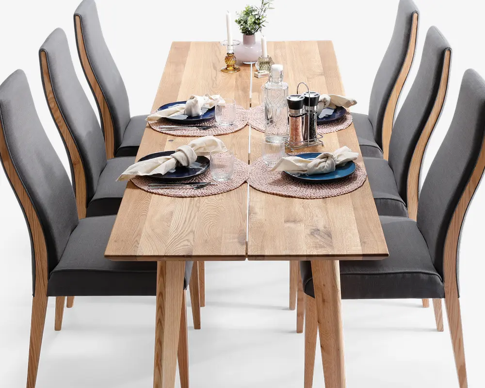 Rektangulært spisebord i træ med naturfinish, omgivet af seks højryggede stole polstret med mørkegråt stof. Bordet er dækket til fire personer med runde flettede dækkeservietter, blå og hvide tallerkener, pænt foldede servietter med servietringe og klart glas. Midten af bordet har en lille potteplante, to hvide lysestager og et sæt salt- og peberbøsser.