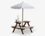 Picnicbord i træ med fastmonterede bænke, designet til børn. Bordet og bænkene har hvide hynder med tynde, mørkeblå striber. Et stort, matchende hvidt og mørkeblåt stribet parasol giver skygge over bordet. På bordpladen står der klare plastikflasker, kopper og et lille fad, hvilket antyder en opsætning til udendørs spisning eller leg.
