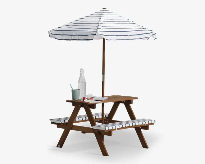 Picnicbord i træ med fastmonterede bænke, designet til børn. Bordet og bænkene har hvide hynder med tynde, mørkeblå striber. Et stort, matchende hvidt og mørkeblåt stribet parasol giver skygge over bordet. På bordpladen står der klare plastikflasker, kopper og et lille fad, hvilket antyder en opsætning til udendørs spisning eller leg.