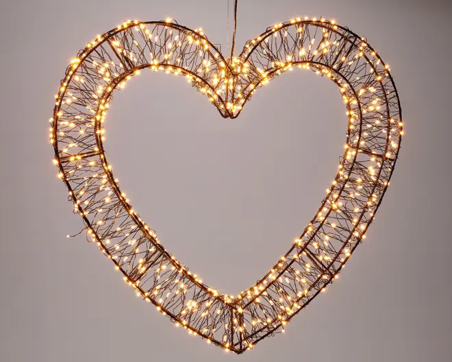 En dekorativ ramme formet som et hjerte, lavet af sammenflettede grene og omviklet med mange små, varm-hvide LED-lyskæder. Lysene er jævnt fordelt langs strukturen og skaber en glødende, elegant kontur. Rammen er let og designet til ophængning, hvilket gør den velegnet til festlig eller romantisk udsmykning.