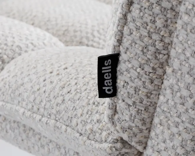 Nærbillede af et lyst beige, tekstureret stof med vævet mønster, der viser detaljeret syning og et sort stofmærke med mærket 'daells' i hvide bogstaver. Stoffet ser blødt og fyldigt ud, hvilket indikerer et behageligt og slidstærkt polstringsmateriale.