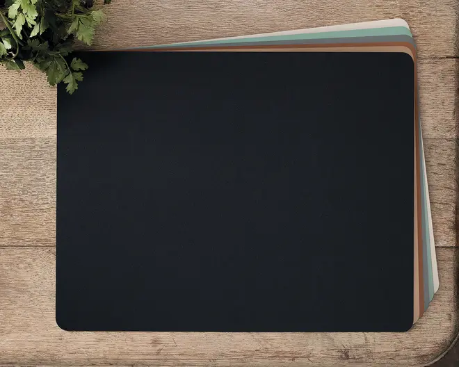 Sæt af rektangulære dækkeservietter med afrundede hjørner, vist i en stak hvor den øverste er ensfarvet sort og de øvrige har afdæmpede farver som brun, blågrøn og lysegrå. Dækkeservietterne har en glat, mat overflade og ser ud til at være lavet af et fleksibelt materiale, der beskytter bordet.