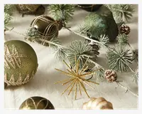 Et udvalg af julepynt med grønne og gyldne kugler med glitrende mønstre, en gylden stjerneformet pynt, frostede kunstige grangrene og små kogler. Kuglerne har både matte og blanke overflader, og grangrenene er dækket af kunstig sne for et festligt, vinterligt udtryk.