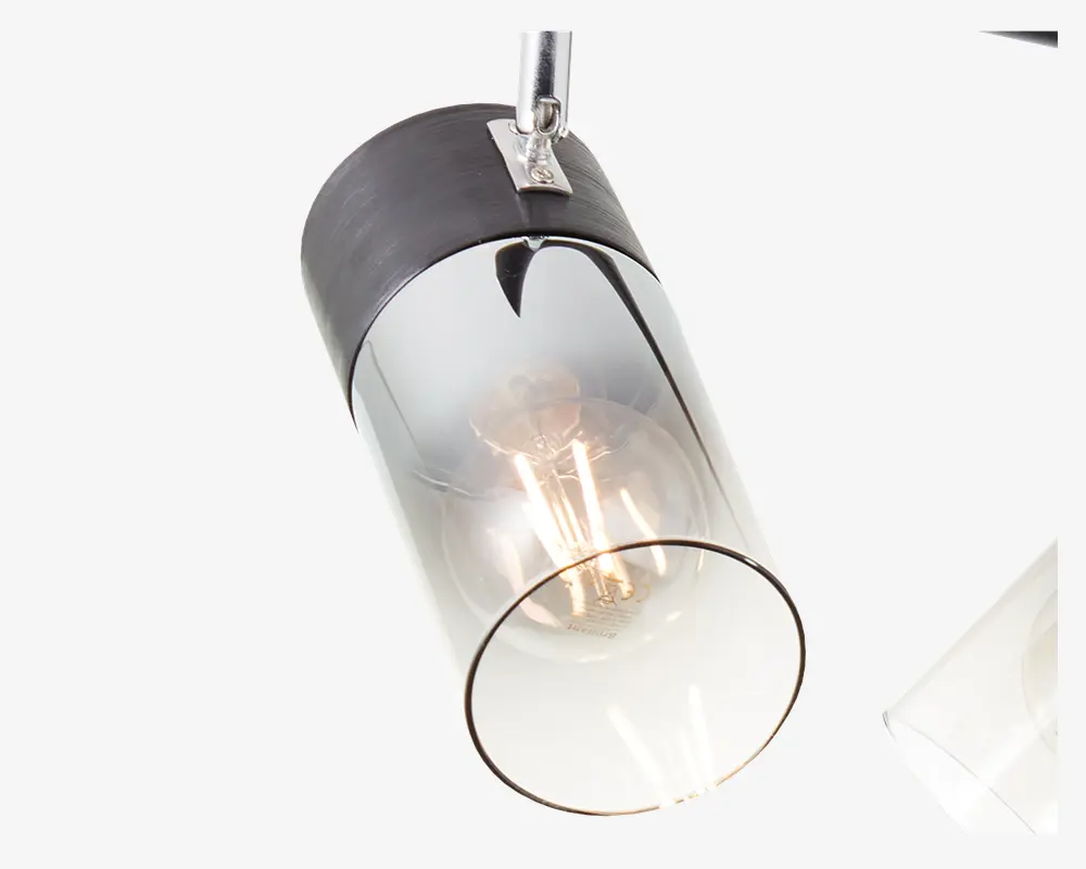 Et nærbillede af en moderne lampe med en cylindrisk glasskærm, der har en diskret overgang fra klart glas i bunden til røgfarvet glas i toppen. Lampen har en sort metalhætte og en synlig glødepære indeni, hvilket giver et industrielt, men elegant udtryk. Glasset er glat og gennemsigtigt, så pæren og dens varme skær tydeligt kan ses.