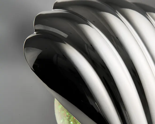 Nærbillede af en blank, sort, rillet overflade med et buet, muslingelignende design. Overfladen er meget reflekterende og viser glatte, flydende linjer samt et elegant, moderne udtryk. Basen har en diskret iriserende eller glitrende effekt.