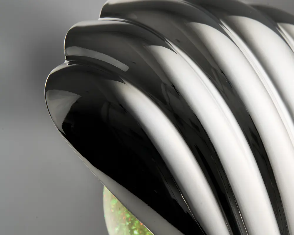 Nærbillede af en blank, sort, rillet overflade med et buet, muslingelignende design. Overfladen er meget reflekterende og viser glatte, flydende linjer samt et elegant, moderne udtryk. Basen har en diskret iriserende eller glitrende effekt.