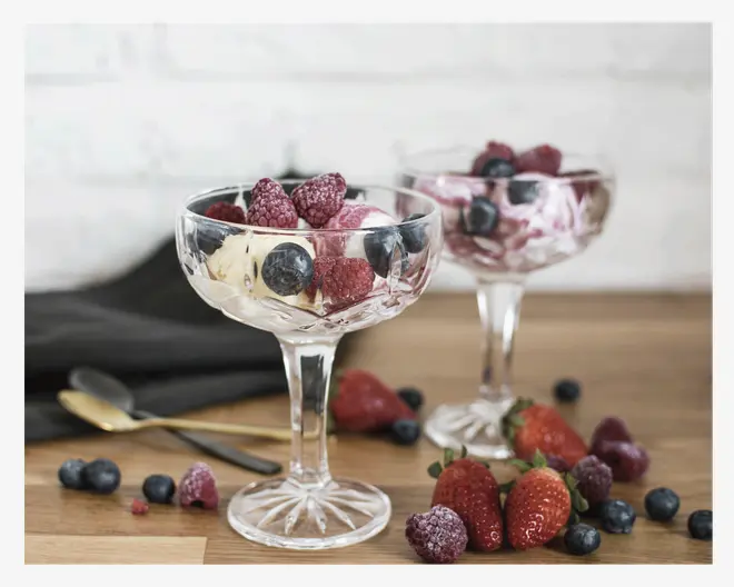 To elegante dessertglas fyldt med kugler af is, toppet med en blanding af frosne hindbær og blåbær. Friske jordbær, blåbær og hindbær ligger spredt på træbordet omkring glassene. Glassene har en klassisk, facetslebet stilk, og der ligger guldfarvede og sølvfarvede dessertskeer ved siden af. Baggrunden viser en hvid murstensvæg og en mørk stofserviet.