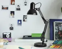 En mat sort justerbar skrivebordslampe med rund fod og leddelt arm, der giver fleksibel placering. Lampen har et klassisk, industrielt design med en kuppelformet skærm, der leder lyset nedad. Den står på et hvidt skrivebord omgivet af kontorartikler, farveblyanter og bøger, hvilket gør den velegnet til studie- eller arbejdsmiljøer.