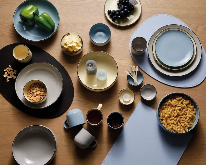 Et koordineret sæt keramikservice i afdæmpede nuancer af blå, beige og creme, bestående af tallerkener, skåle, krus og kopper. Sættet har både runde og ovale former, hvor nogle dele har en kontrastfarvet kant. Service er arrangeret på en træbordplade med dækkeservietter i sort og lyseblå. Nogle skåle og tallerkener indeholder madvarer som grønne peberfrugter, vindruer, pasta, peanuts og chips, mens andre er tomme eller indeholder stearinlys. Designet er moderne og minimalistisk, velegnet til både hverdags- og festbrug.