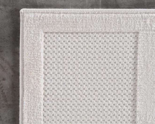 Nærbillede af et rektangulært tekstilprodukt med et tekstureret, vævet midterpanel og en bred, ensfarvet kant. Stoffet er råhvidt eller lysebeige, og kanten har en lidt højere luv end midten, hvilket giver en diskret kontrast i tekstur. Syningen langs kanten er pæn og forstærker kanten.
