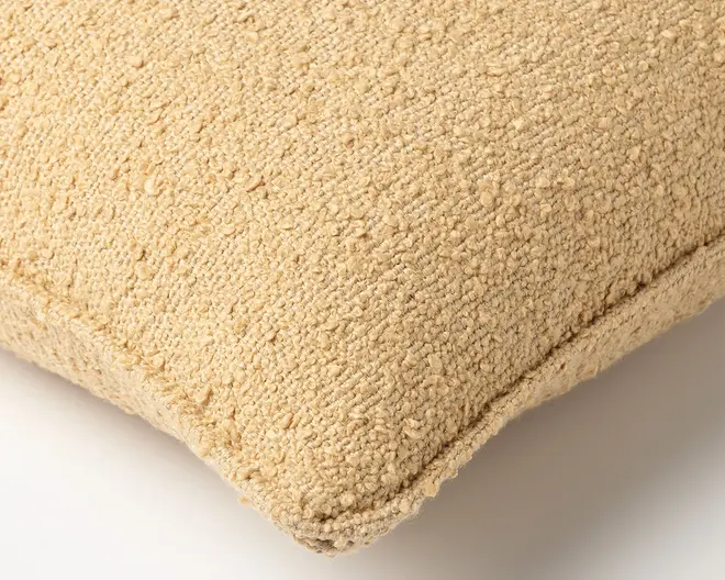 Nærbillede af en lysebeige, tekstureret bouclé-pude med afrundet hjørne og synlig søm.
