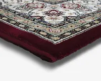 Nærbillede af et tykt, blødt tæppehjørne med en dyb bordeaux kant og et detaljeret, ornamenteret mønster med creme, grønne og røde nuancer. Tæppet har en blød, tæt luv og et klassisk design med blomster- og geometriske motiver.