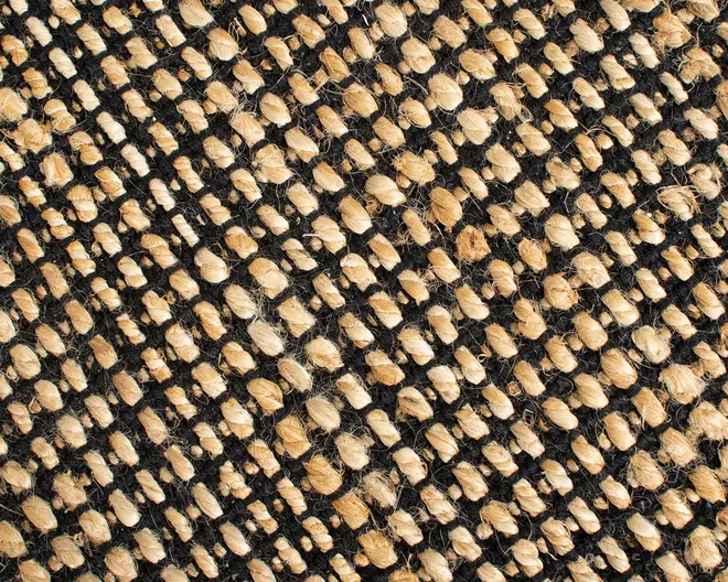 Nærbillede af en vævet tekstur med tykke, naturlige beige fibre, der er flettet sammen med sorte tråde. Vævet er groft og kraftigt, hvilket giver et rustikt og slidstærkt udtryk. Fibrene har et let ujævnt, organisk udseende med synlige frynser, hvilket indikerer et håndlavet eller naturligt materiale. Mønsteret er diagonalt, hvor de beige fibre danner markante forhøjede rækker mod den sorte baggrund.