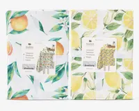 To sammenfoldede stofgenstande med farverige frugt-motiver. Den til venstre har appelsiner og grønne blade på hvid baggrund, samt små hvide blomster. Den til højre viser gule citroner, citronskiver og grønne blade, også på hvid baggrund. Begge er pænt pakket ind med et hvidt bånd og et produktmærke, der viser et eksempel på det fulde design.