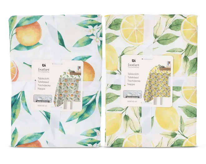 To sammenfoldede stofgenstande med farverige frugt-motiver. Den til venstre har appelsiner og grønne blade på hvid baggrund, samt små hvide blomster. Den til højre viser gule citroner, citronskiver og grønne blade, også på hvid baggrund. Begge er pænt pakket ind med et hvidt bånd og et produktmærke, der viser et eksempel på det fulde design.