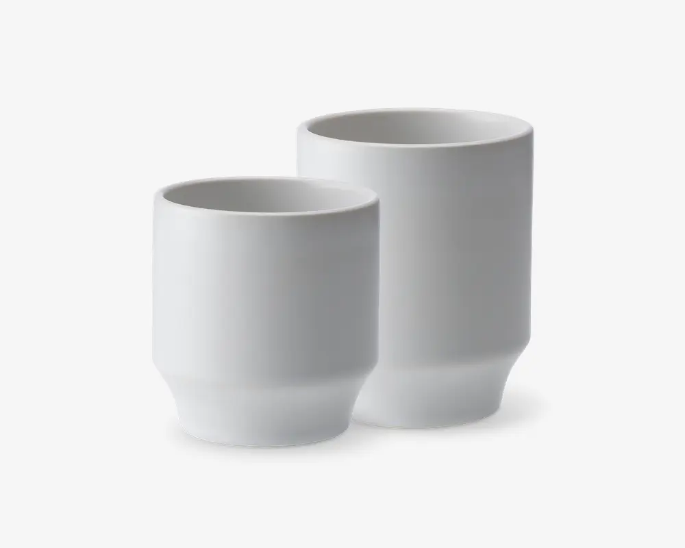 To minimalistiske, cylindriske hvide kopper med mat finish. Begge kopper har en let tilspidset bund og en glat, ensfarvet overflade, hvilket giver dem et moderne og elegant udtryk. Kopperne har forskellig højde, hvor den ene er højere end den anden, men de har samme enkle design.