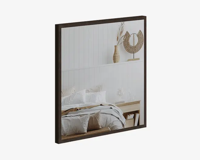 Firkantet spejl med en mørk, slank træramme. Spejlet reflekterer et hyggeligt soveværelse med en seng med hvidt sengetøj, en træbænk for enden af sengen og en hylde over sengen med dekorative genstande som en fletvase med tørrede blomster og en cirkulær rattan-skulptur. Stilen er moderne og minimalistisk med naturlige, jordnære nuancer.