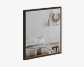 Firkantet spejl med en mørk, slank træramme. Spejlet reflekterer et hyggeligt soveværelse med en seng med hvidt sengetøj, en træbænk for enden af sengen og en hylde over sengen med dekorative genstande som en fletvase med tørrede blomster og en cirkulær rattan-skulptur. Stilen er moderne og minimalistisk med naturlige, jordnære nuancer.