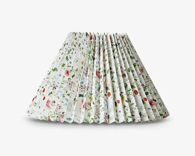Plisseret lampeskærm med hvid baggrund og et fint blomstermønster i forskellige farver, herunder grønne blade samt røde, lilla og orange blomster. Plisseringen giver en tekstureret og tredimensionel effekt, og blomstermotivet er jævnt fordelt over hele overfladen.