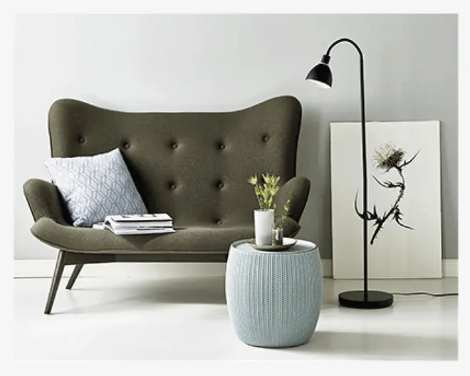 Et moderne stuearrangement med en mørke olivengrøn, sofa i retrostil med buet ryglæn og knapdetaljer. Sofaen har skrå træben og er pyntet med en lysegrå pyntepude med mønster samt en stak bøger. Foran sofaen står en rund, lyseblå strikket puf, der bruges som sofabord og har en lille hvid vase med grønne grene og et lys på toppen. Til højre står en sort gulvlampe med buet hals ved siden af et minimalistisk botanisk kunsttryk, der hviler op ad en lysegrå væg.