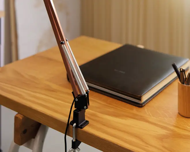Et nærbillede af et skrivebord i træ med en solid, justerbar arm monteret med klemme og synlige metalskruer, sandsynligvis til en lampe eller mikrofon. Bordpladen har en glat overflade med naturlig træstruktur. På bordet ligger en stor, lukket sort notesbog eller kalender med tykke sider samt en træholder med flere kuglepenne eller blyanter. Opsætningen giver et indtryk af en minimalistisk og organiseret arbejdsplads.