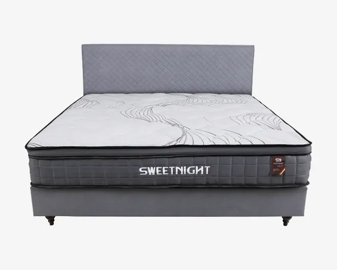 En moderne seng med et quiltet, lysegråt sengegærde og en tyk, blød madras med hvid top og diskrete grå bølgemønstre. Madrassen har en mørkegrå side med ternet mønster og mærket 'SWEETNIGHT' tydeligt vist med hvide bogstaver. Sengebunden er betrukket med matchende gråt stof og står på korte, solide ben.
