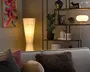 Høj, konisk gulvlampe med plisseret hvid skærm, der lyser varmt ved siden af en moderne reol og sofa.