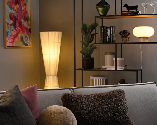 Høj, konisk gulvlampe med plisseret hvid skærm, der lyser varmt ved siden af en moderne reol og sofa.