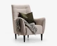 Moderne lænestol betrukket med lysebeige stof og en let buet ryg- og siddeflade. Stolen har slanke, skråtstillede sorte ben og leveres med to dekorative grønne puder. Et tekstureret, frynset gråt tæppe er lagt over den ene armlæn, hvilket giver et hyggeligt udtryk.