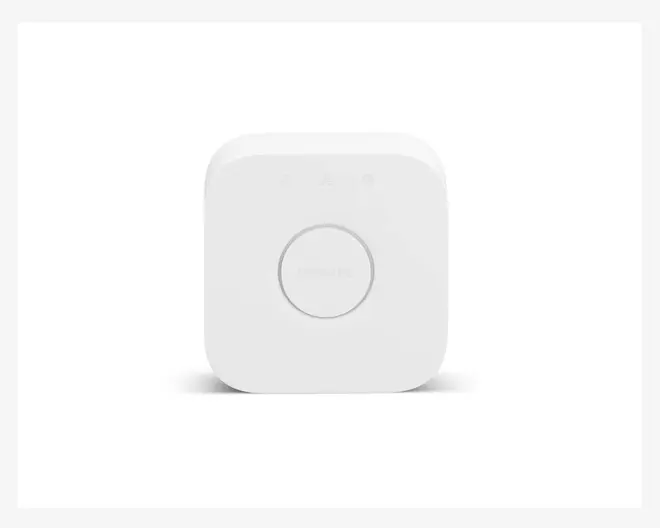 En lille, hvid, firkantet enhed med afrundede hjørner. Den har en stor, cirkelformet knap i midten med diskret tekst på knappen. Over knappen er der tre små indikatorlys. Designet er minimalistisk og moderne med en glat, mat overflade.