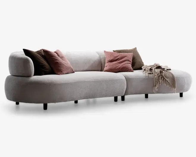 Buet, moderne sofa betrukket med lysebeige stof med lav ryg og afrundede kanter. Sofaen har fem bløde pyntepuder i brune og støvede rosa nuancer samt et tekstureret beige tæppe lagt over den ene arm. Sofaen står på korte, diskrete sorte ben, hvilket giver et svævende udtryk.