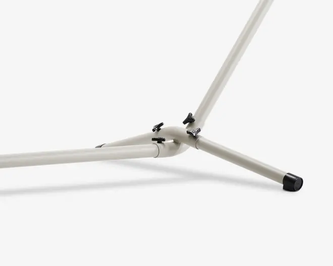Nærbillede af en robust, lysegrå metalramme med tre rørformede ben, der samles i et centralt knudepunkt. Benene er fastgjort med sorte låsepinde, og det ene ben har en sort gummifod for stabilitet og beskyttelse af gulvet. Metallet har en glat, mat overflade.