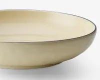 En lav, rund skål med en glat, blank cremefarvet overflade. Skålens kant er diskret markeret med en tynd, mørk streg, som giver en elegant kontrast til den ensartede farve. Skålen har blødt skrånende sider og et minimalistisk, moderne design.